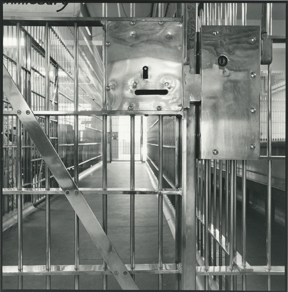 Black-and-white photograph focused on the lock on a door with bars leading to a hallway of prison cells