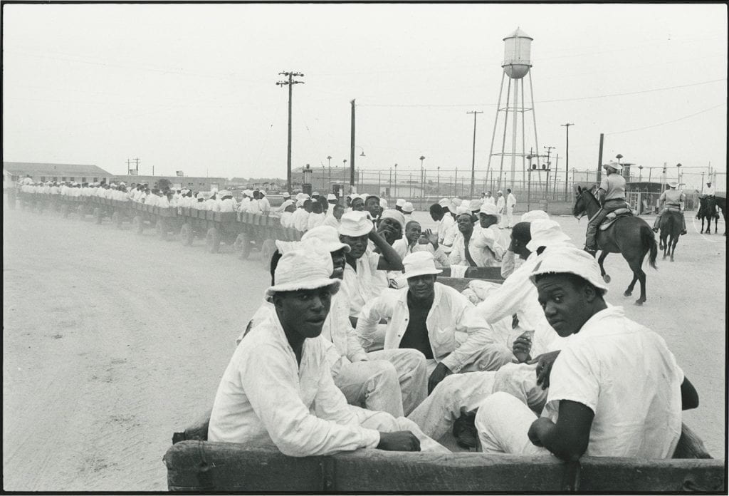 Black-and-white photograph of dozens of Black men being transported in a long train of wooden carts, three prison guards on horseback nearby