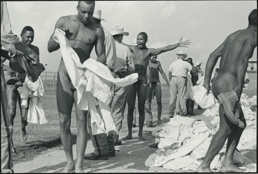 Black-and-white photograph of nude Black men being searched by white prison guards