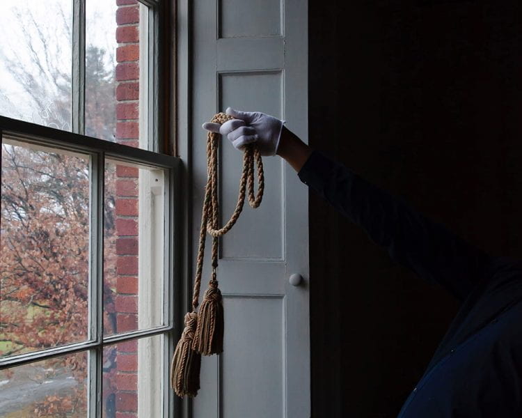 figure, leaning backwards so as to have their torso cloaked in shadow, standing by a window, holding a thick tasseled cord with a white-gloved hand