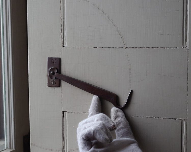 Photograph of a white gloved hand lifting a hook lock on a white door