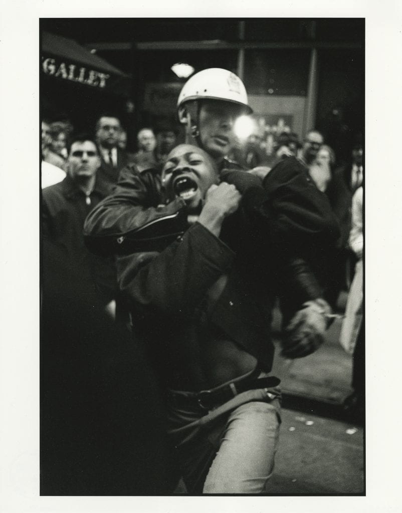 Black and White photograph centered on White, helmeted police officer as he drags a Black teenager by the neck in a chokehold, amidst a grown of mostly White onlookers in a crowded street scene